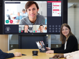 A modern video conference in progress, displaying a man on the main screen with additional participants in smaller windows, while two individuals collaborate in a conference room setting.
